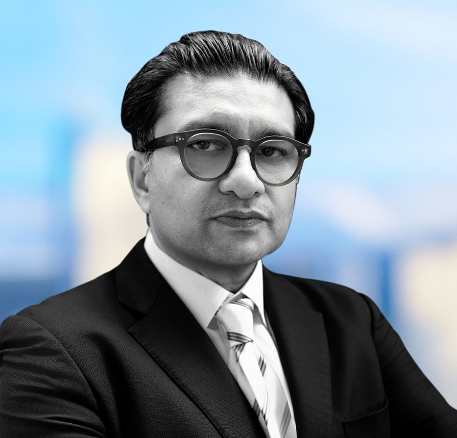 Abbas - ARM Investments - ARM Investments Professional headshot of a man in a suit and glasses against a blue blurred cityscape backdrop.
